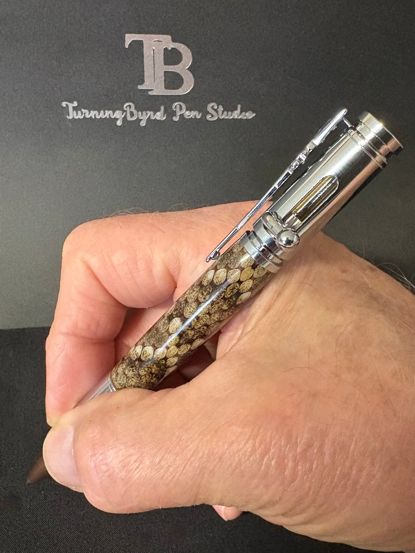 Hand holding a pen made with rattlesnake skin with 'TB Turning Byrd Pen Studio' branding in the background