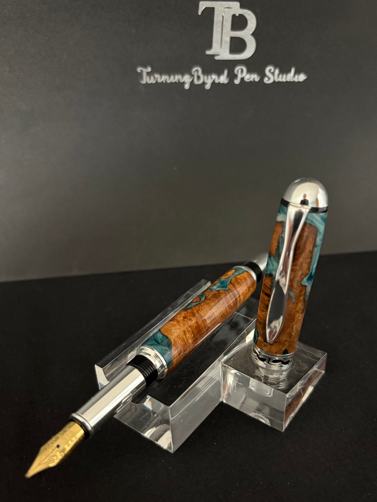 Cherry Burl Wood pen with green and white resin on a clear stand against a gray background with 'TB Turning Byrd Pen Studio' logo.