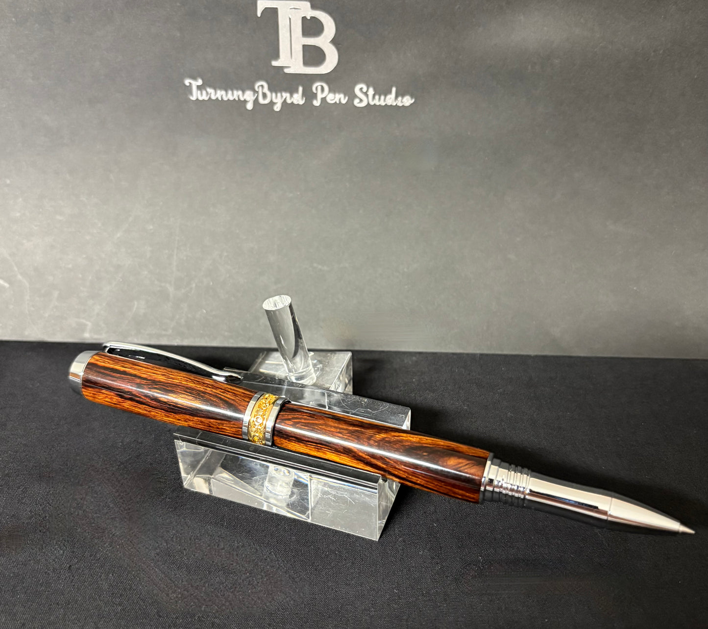 Desert Ironwood pen with silver accents on a clear stand against a gray background with 'Turning Byrd Pen Studios' logo.