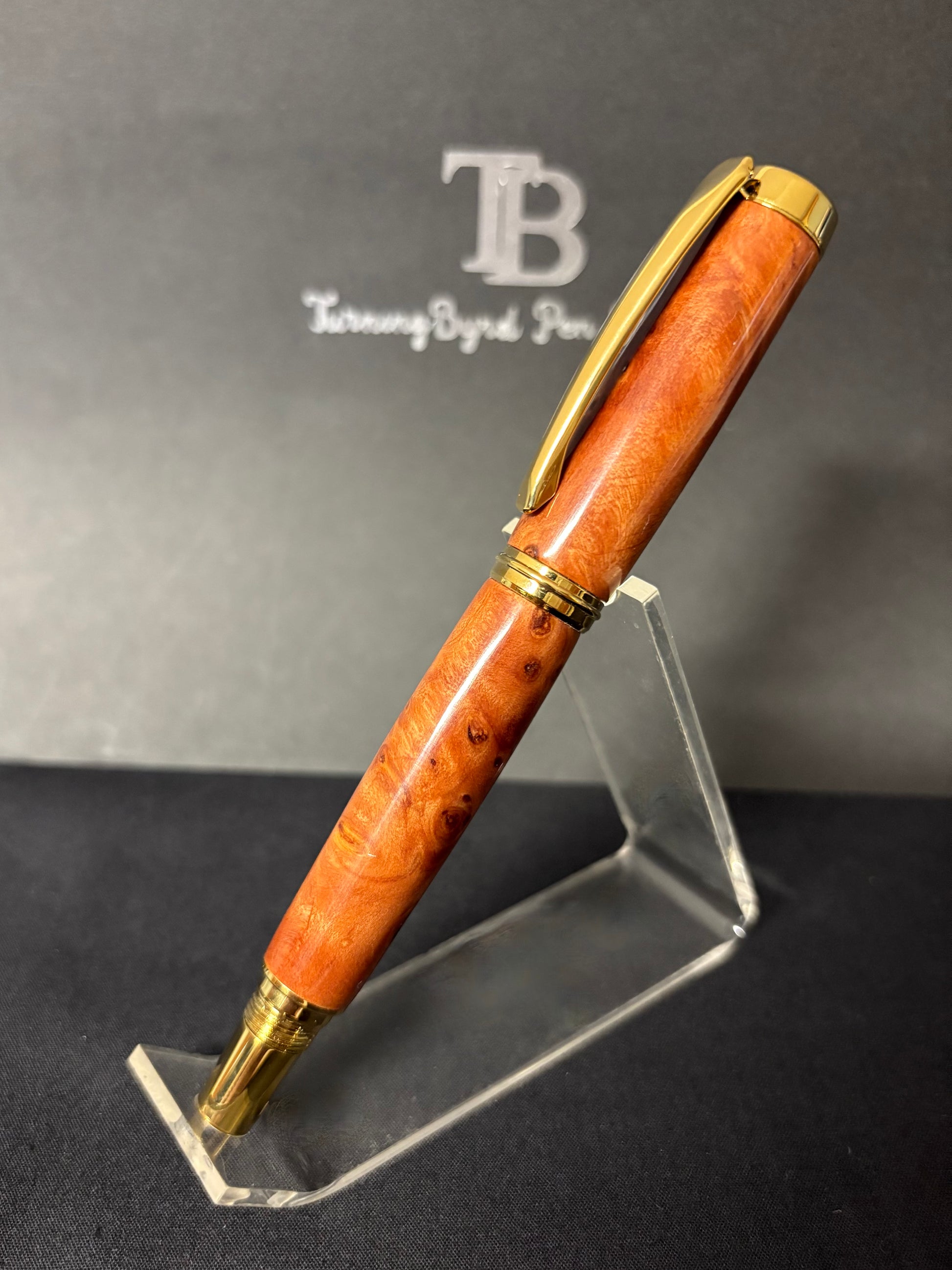Redwood Burl pen with gold accents on a clear stand against a dark background with a brand logo.