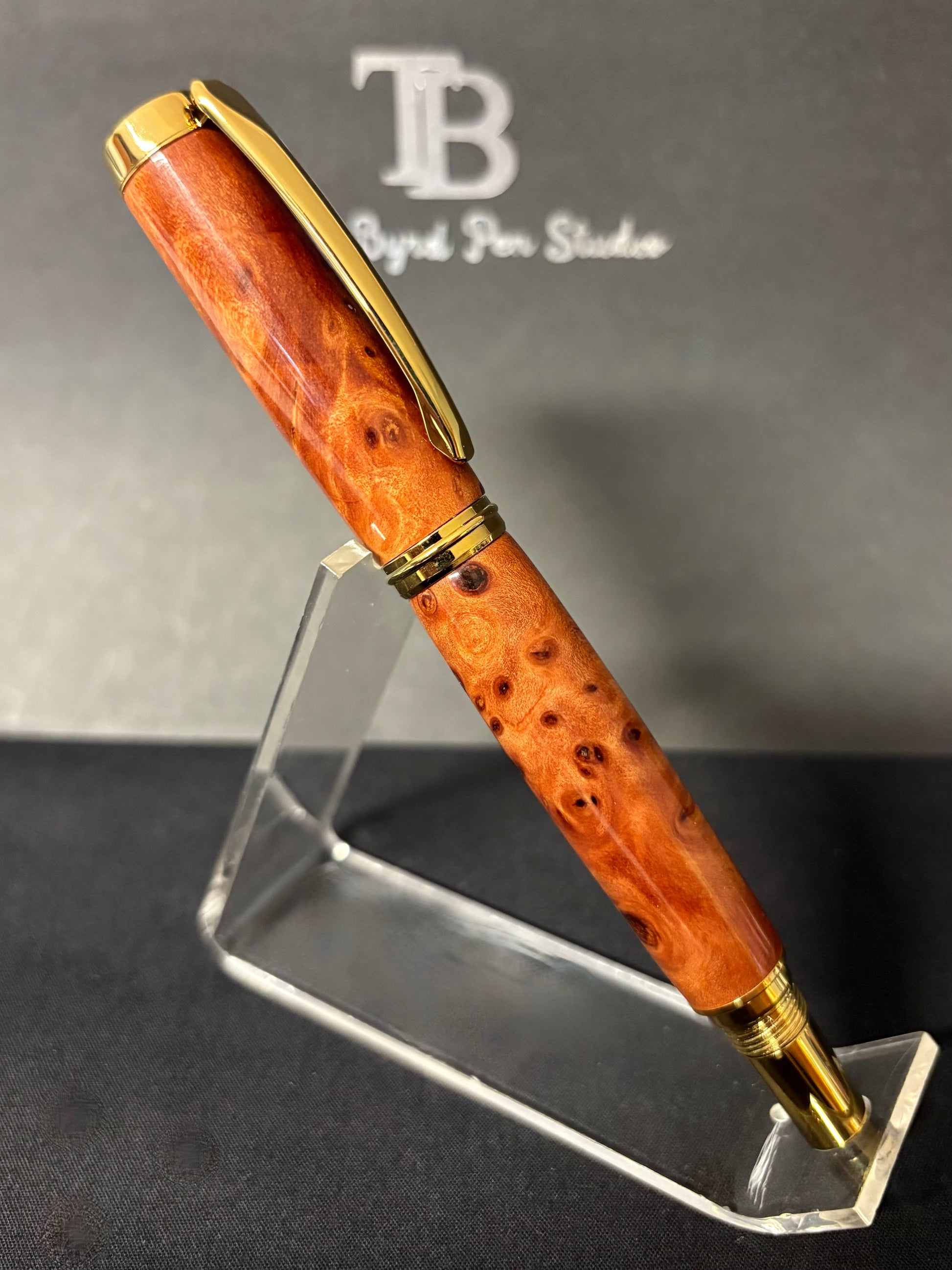 Redwood Burl pen with gold accents on a clear stand against a branded background