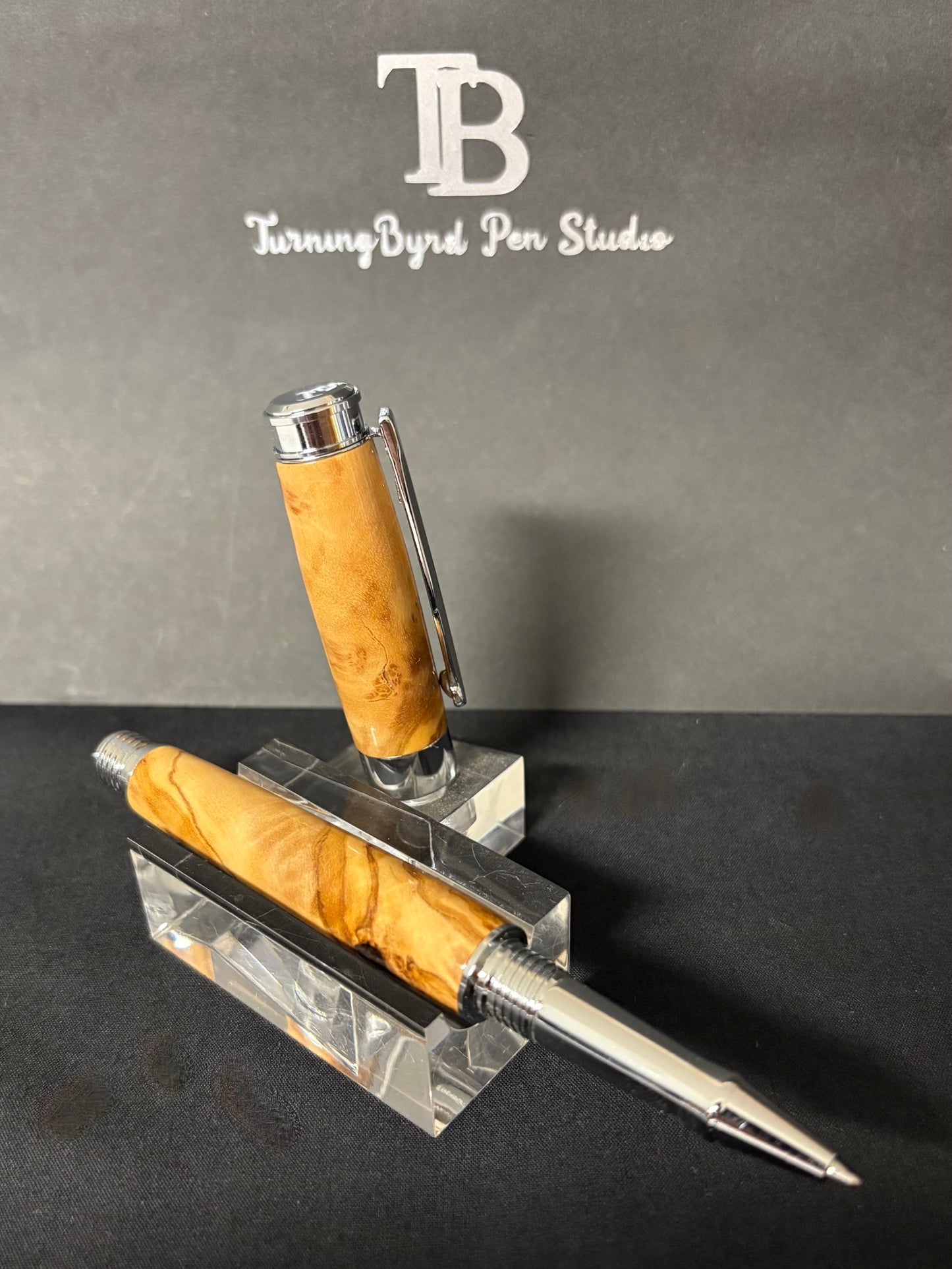 Olive Wood rollerball pen with metal clip  on a stand, branded 'TurningByrd Pen Studio'.