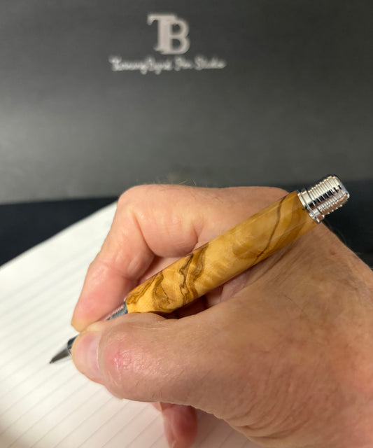 Hand holding a Majestic Olive Wood pen with a wooden grip and silver clip against a black background with 'TB' branding.