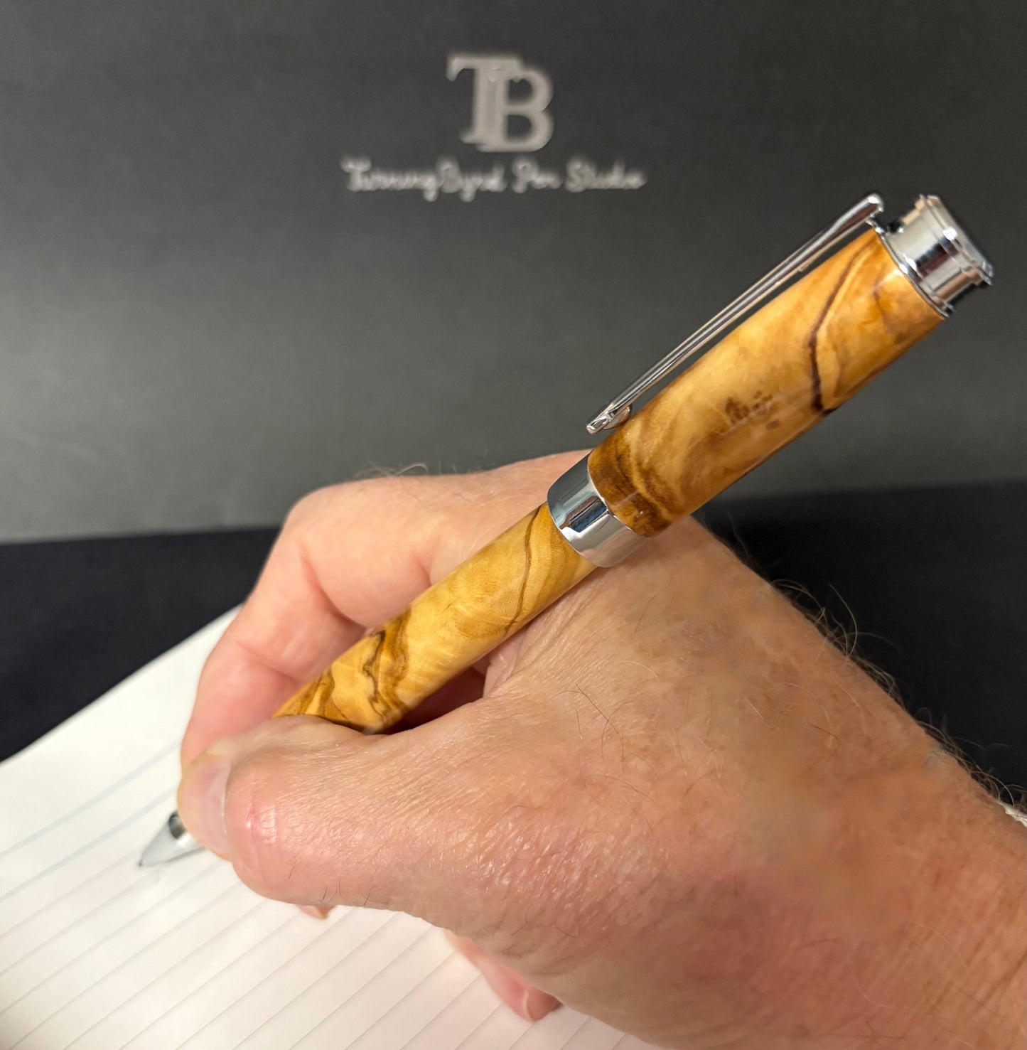 Hand holding a Postable Olive Wood Rollerball  pen with silver clip, blurred TB logo in the background