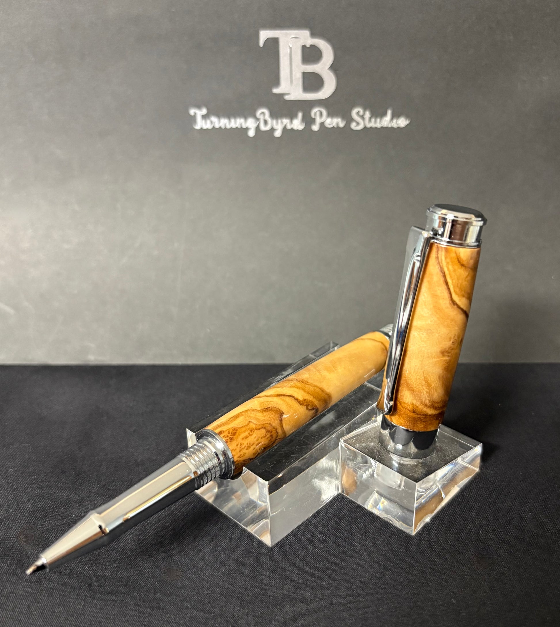 Olive Wood pen with silver accents on a clear stand against a gray background with 'Turning Byrd Pen Studio' branding.